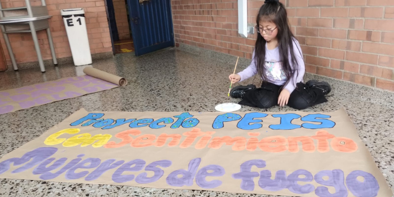 Niña en el suelo mostrando una pancarta que dice: Proyecto PEIS ConSentimiento Mujeres de Fuego