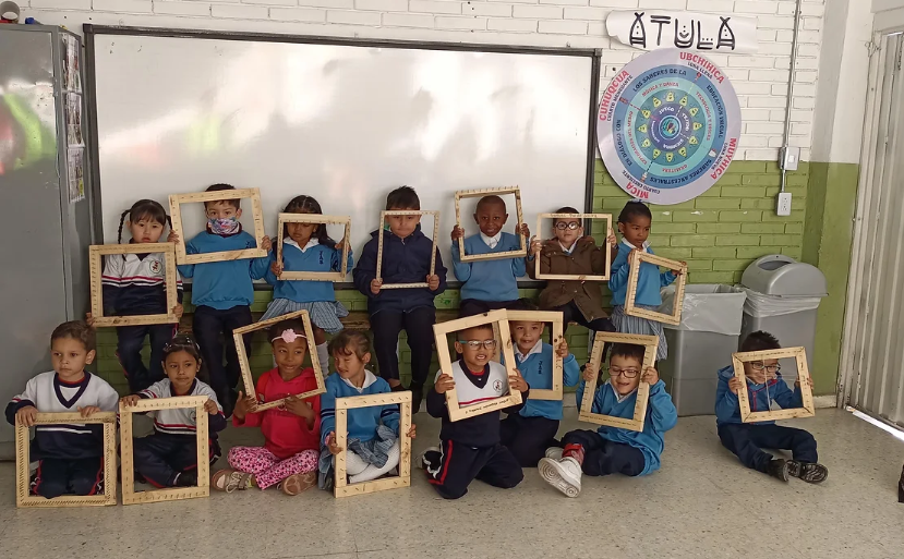 Foto de niños y niñas en una salón de clases, sosteniendo un marco de madera cada uno